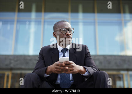 Low angle portrait of contemporary African-American businessman using smartphone sitting on steps outside office building during break and looking awa Stock Photo
