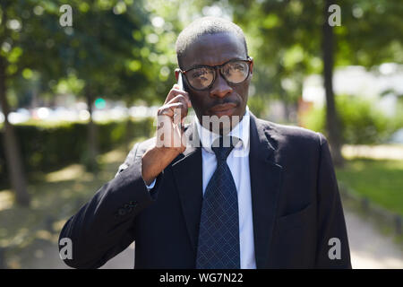 Waist up portrait of successful African-American man speaking by smartphone while walking outdoors in park lane, copy space Stock Photo