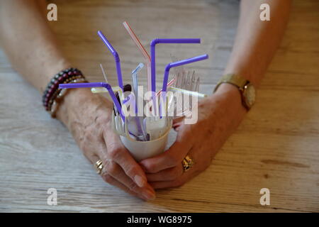 Senior female hands on wooden table holds white plastic cup with plastic disposable drinking straws, cutlery like spoons, forks, knives inside closeup Stock Photo