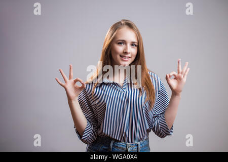 A truly charming red haired girl with two hands showing OK sign on gray background with copy space for text. Good mood, luck, happiness and emotion co Stock Photo