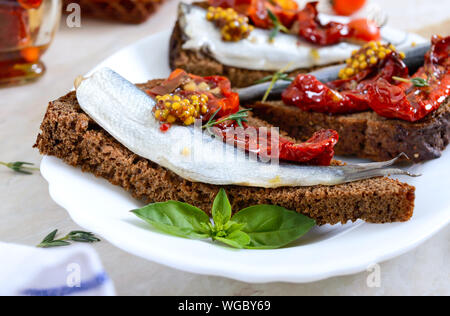 Tasty smorrebrod on a white plate. Sandwiches with black rye bread, sun-dried tomatoes, salted anchovies, mustard. Stock Photo