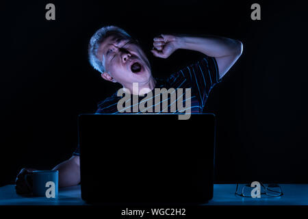 Tired middle age Asian male yawning while working on his computer in his home office at night Stock Photo