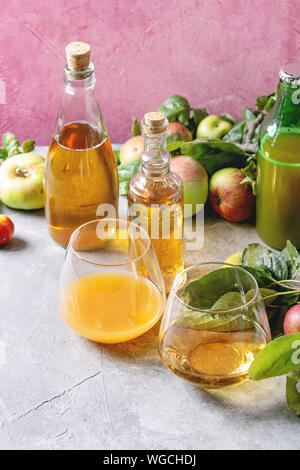 Variety of apple drinks. Bottles ang glasses of apple juice, vinegar and cider with garden apples with leaves and branches on table with pink backgrou Stock Photo