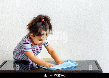 Cute brunette child helping her mom washing modern design garden furniture, glass table on home terrace. Little girl make cleaning with water and blue towel. Stock Photo