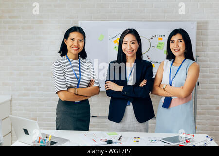 Smiling Asian young business women in casual wear standing in line with arms folded in meeting room. Row of Business men and women looking at camera Stock Photo