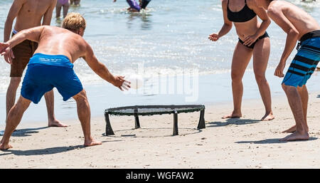 Four people playing a black and white spike ball game on the sand at the beach next to the ocean. Stock Photo