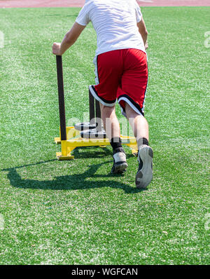 A teenage boy is pushing a yellow sled with weights on it during running camp on a green turf field. Stock Photo