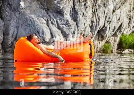 The tourist fell asleep in an orange air sofa or an inflatable lounge floating on the river. Rafting on the wild river on an inflatable lounger, while Stock Photo