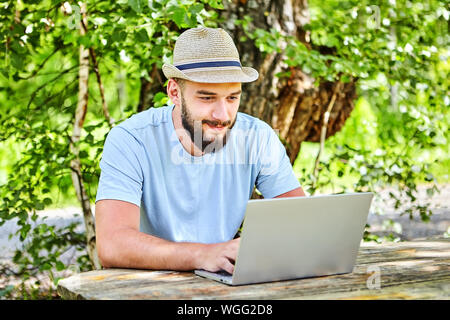 A bearded young Caucasian man, with a wicker hat on his head, looks at the laptop screen with a pleased expression on his face. A student or businessm Stock Photo