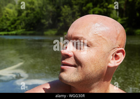 A bald man is resting in the wild in the ecosystem of a forest river, he smiles when a moth sits on his nose. Ecotourism in the nature reserves of Rus Stock Photo