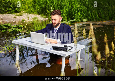 The table is in the river, behind it is a businessman, a white collar, or an office worker dressed in a suit, he is waist-deep in water, and works on Stock Photo