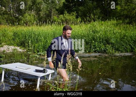 A man in a suit, but without pants, leaves a workplace located in a river ecosystem in the wild. A businessman steps knee-deep in the water, leaving b Stock Photo