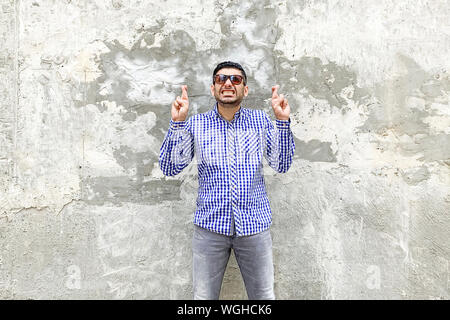 Portrait of hopeful handsome bearded young man in checkered blue shirt and sunglasses standing against concrete gray wall. crossed finger, clenching t Stock Photo