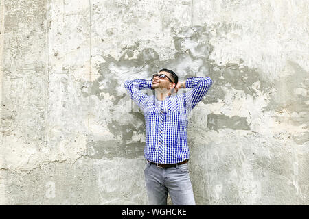 Portrait of happy handsome bearded young man in checkered blue shirt and sunglasses standing against concrete gray wall. holding his head and looking Stock Photo