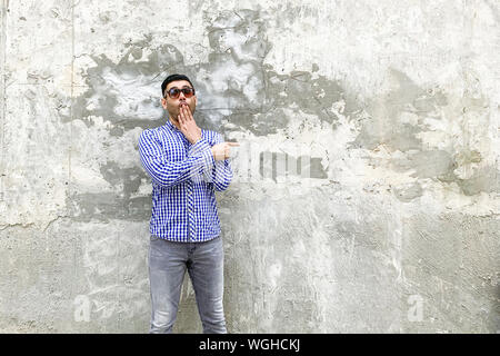 Portrait of shocked bearded young man in checkered blue shirt, sunglasses standing against concrete gray wall. pointing at background empty copyspace Stock Photo