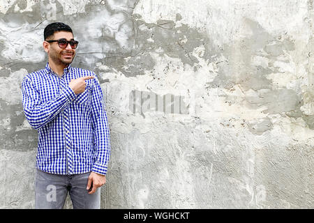 Portrait of happy handsome bearded young man in checkered blue shirt and sunglasses standing against concrete gray wall. pointing at background empty Stock Photo