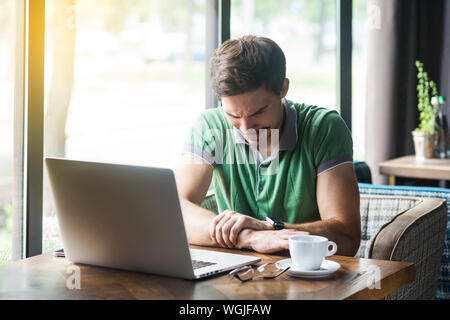 Hand pain. Young businessman in green t-shirt sitting and feeling ache on his hand after too much working and typing on laptop. business and freelanci Stock Photo