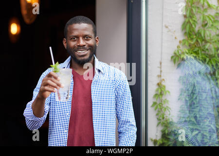 Waist up portrait of smiling African-American man holding cold drink outdoors while posing by cafe, copy space Stock Photo