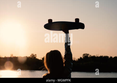 Silhouetted Woman posing with her hands holding skateboard raised up in the sunset sky, summer farewell concept. Stock Photo