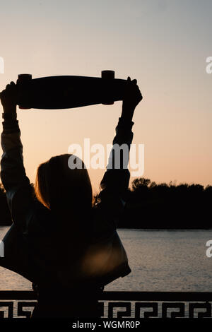 Woman posing with her hands holding skateboard raised up in the sunset sky, summer farewell concept. Vertical image. Stock Photo