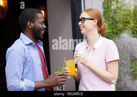 Waist up portrait of smiling young woman talking to African man outdoors, both holding cold refreshing drinks, copy space