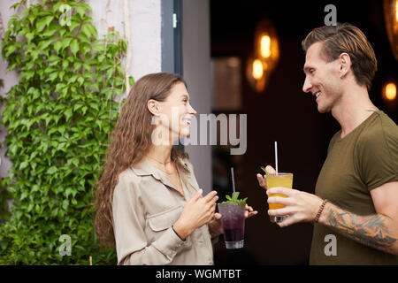 Side view portrait of smiling young woman talking to man outdoors standing by cafe door, both holding cold refreshing drinks, copy space Stock Photo