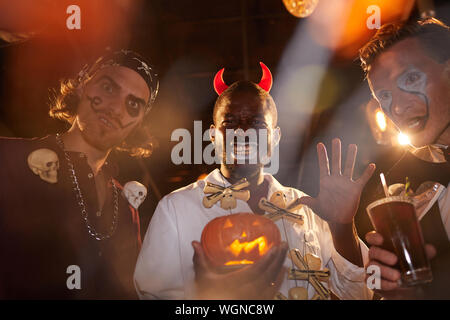 Group of adult men wearing Halloween costumes posing during party in club, focus on African-American man holding pumpkin in center Stock Photo
