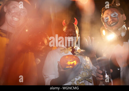 Low angle view at group of adult men wearing Halloween costumes posing during party in club, focus on African-American man holding pumpkin in center Stock Photo