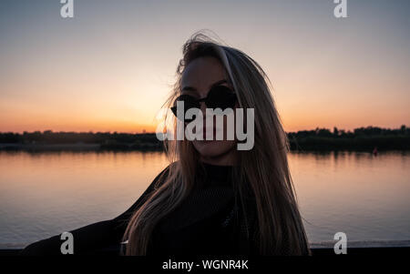 Thoughtful woman in her 30s posing in front of lake sunset in trendy sunglasses Stock Photo