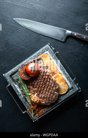 Grilled steak with knife and fork carved on black stone slate. Steak on a hot marble stone. Copy space, dark background, food fashion photo Stock Photo