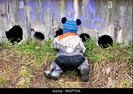 A little boy of two or three years old is kneeling to look through holes in the wall and feed the kittens Stock Photo