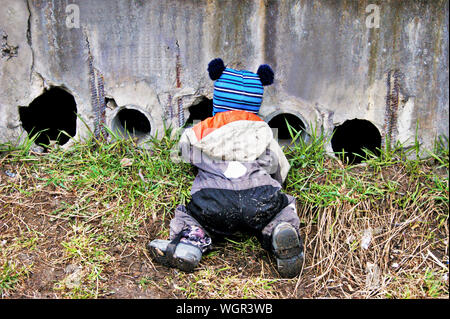 A little boy of two or three years old is kneeling to look through holes in the wall and feed the kittens Stock Photo