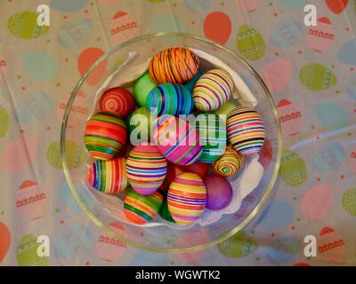 A collection of eggs dyed for easter in a glass bowl which is sitting on a plastic table cloth imprinted with an easter theme. Stock Photo