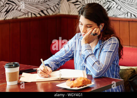Young bored woman sitting at cafe table, listening to lection in earbuds and writing down important thoughts Stock Photo