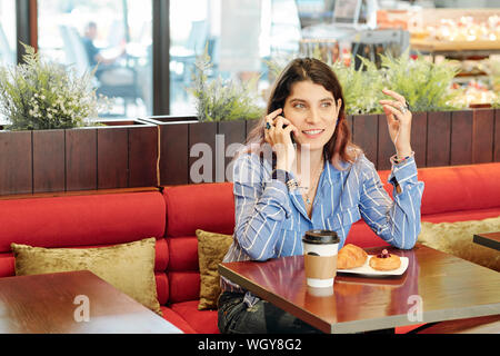 Portrait of young businesswoman sitting at table in cafe and discussing important issues on smartphone with colleagues Stock Photo