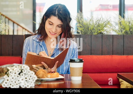 Pretty smiling young woman sitting at table in cafe with coffee and couquet of flowers and taking notes in diary Stock Photo