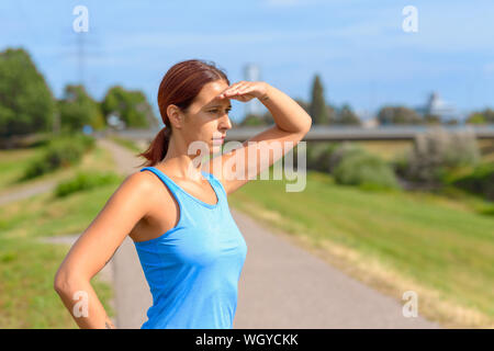 Attractive tanned woman standing watching with her hand to her eyes and a thoughtful serious expression outdoors in summer sunshine Stock Photo