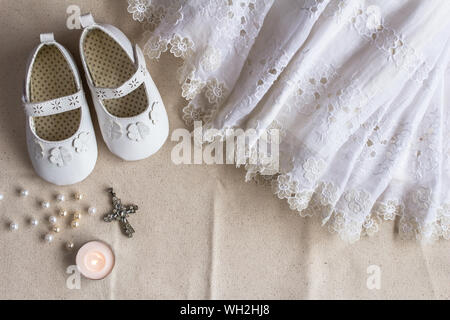 Christening background with baptism dress, shoes, candles and crystal cross pendant on white linen cloth Stock Photo