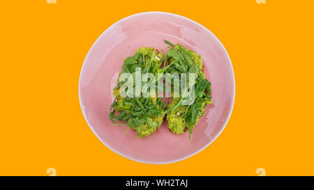 Two slices of mashed avocado toast garnished with fresh rocket or arugula salad set on a pink plate against a bright orange background Stock Photo