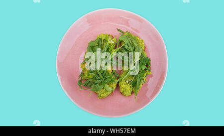 Two slices of mashed avocado toast garnished with fresh rocket or arugula salad set on a pink plate against an aqua blue background Stock Photo