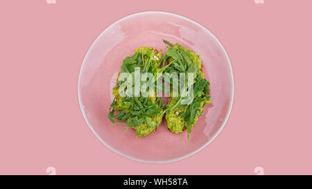 Two slices of mashed avocado toast garnished with fresh rocket or arugula salad set on a pink plate against a pink background, ideal snacks or meals Stock Photo