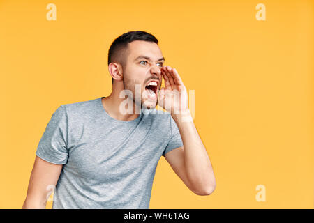 Young angry man shouting and screaming loud to side with hand on mouth and copy space for text over yellow background. Attention, communication emotio Stock Photo