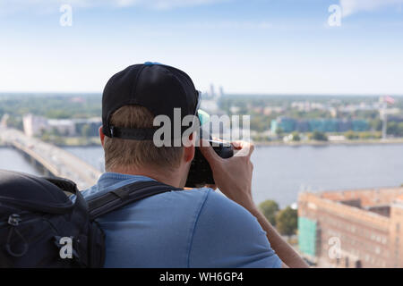 Photographer taking picture above Riga cityscape. Tourist taking picture from view point. Man with backpack, camera and hat. Travel concept. Stock Photo