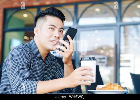 Positive handsome young VIetnamese man having cup of coffee and recodring voice message for smartphone Stock Photo