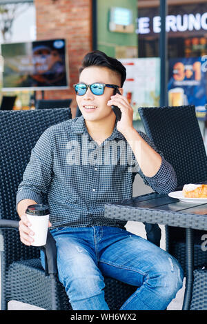 Cheerful young man in sunglasses sitting at cafe table, enjoying tsty breakfast and making phone call Stock Photo