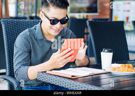 Portrait of positive young man sitting at cafe table, having breakfast and reading news online Stock Photo