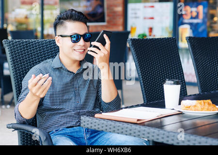 Smiling Asian businessman sitting at cafe table and recording voice message to explain work to colleague Stock Photo