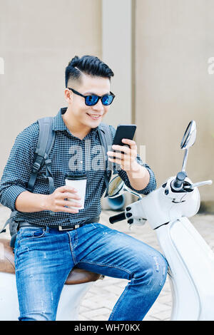 Smiling cool young Asian man sitting on scooter, drinking coffee in disposable cup and reading text messages in his smartphone Stock Photo