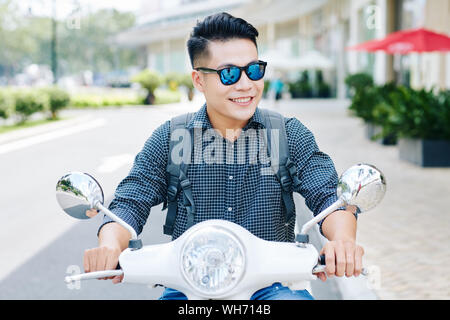 Happy young smiling Vietnamese man with backpack enjoying riding in city streets on scooter Stock Photo