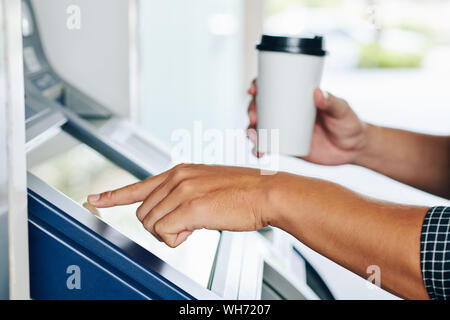 Close-up image of person with cup of coffee in hands entering pin code at ATM machine and withdrawing money from banking account Stock Photo